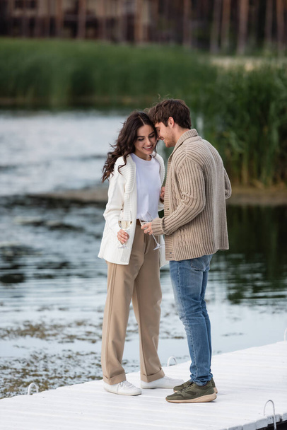 Smiling couple in cardigans holding wine on pier near river  - Foto, imagen