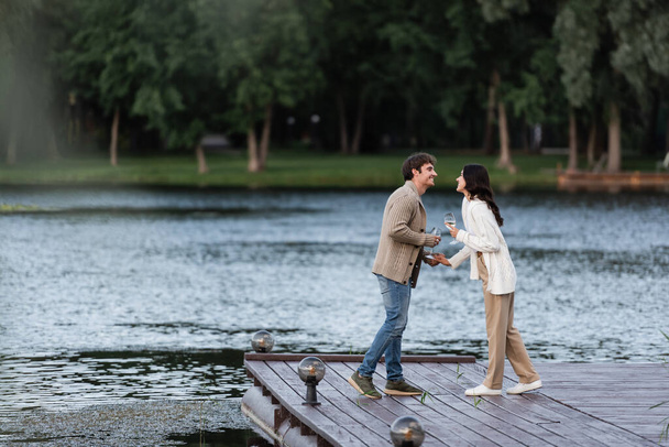 Side view of positive couple in cardigans holding wine on pier near lake  - Photo, Image