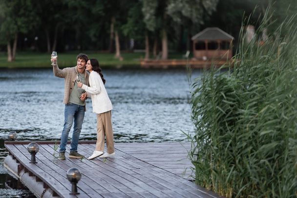 Smiling couple in cardigans holding wine on pier near lake  - Foto, Imagem