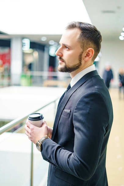 A young bearded man dressed in a business suit at the mall with a cup of coffee. A young man in an elegant business suit strolls through a mall drinking coffee as he goes - Fotoğraf, Görsel