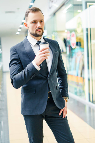 A young bearded man dressed in a business suit at the mall with a cup of coffee. A young man in an elegant business suit strolls through a mall drinking coffee as he goes - Fotó, kép