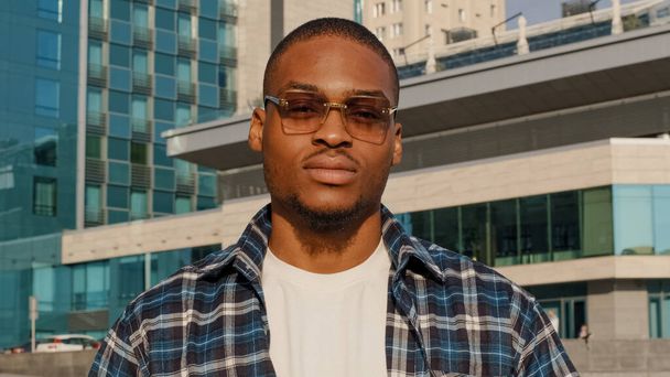 Portrait of man in city african american guy young student man in stylish sunglasses wears plaid shirt looks posing dreaming thinking contemplates turns head looking at camera serious face expression - Φωτογραφία, εικόνα