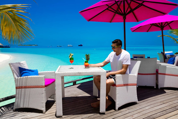 Handsome smiling man is sitting in beach bar on the Maldives sandy beach and holding colorful fruity drink. Luxury travel. Tourism. Maldives landscape. Male model with colorful drink. - Fotografie, Obrázek