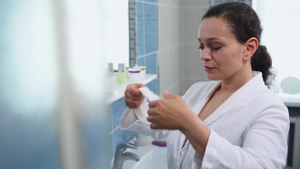 Beautiful middle-aged multi-ethnic woman wearing a white bathrobe, stands in the bathroom and puts on a terry headband before morning beauty procedures. Hygiene. Skin and body care. Daily routine - 映像、動画