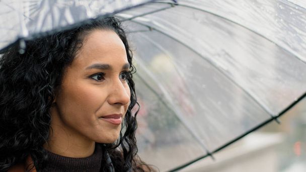 Porträt sorglos attraktive schöne Frau weiblich glücklich hispanischen Mädchen mit langen dunklen lockigen Haaren hält transparenten Regenschirm in der Stadt bei regnerischem Wetter regen lächelnd zahm Blick in die Kamera - Foto, Bild