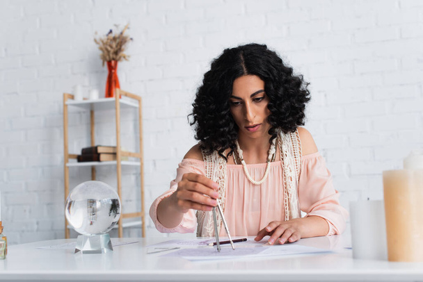 brunette astrologer drawing star charts near crystal ball at home - Foto, Bild