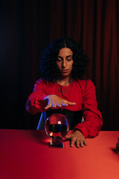 brunette fortune teller with closed eyes holding hand over crystal ball on dark background with red drape - Photo, Image
