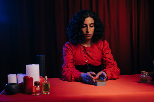 brunette medium sitting near tarot cards and candles on dark background with red drape - Foto, imagen