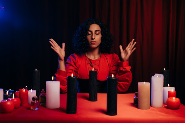 young oracle with closed eyes during spiritual session near burning candles on dark background with red drape - Foto, Imagem