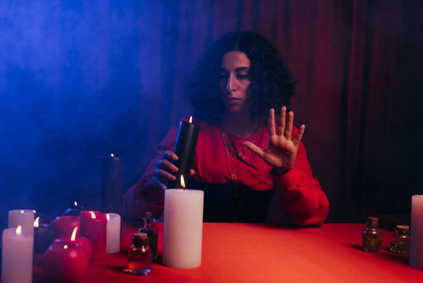 brunette oracle holding burning candle near red table on dark background with blue smoke - Photo, image