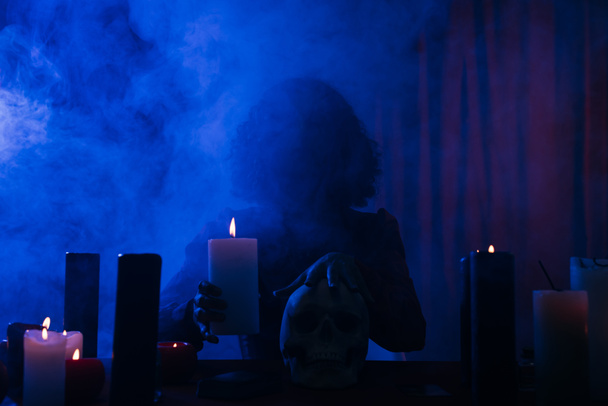 medium holding candle and touching skull during spiritual session in darkness with blue smoke - Φωτογραφία, εικόνα