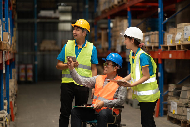 Manager handicapped in wheelchair man employee using tablet check stock work at warehouse. Worker wearing high visibility clothing and hard hat, helmet and checking count up goods boxes for delivery. - Foto, immagini