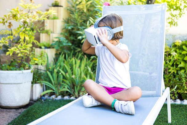 Little girl with virtual reality glasses, holding them with her hands, looking to the left, sitting on a sun lounger in the garden of her house. Metaverse, VR, game, digital and simulation concept. - 写真・画像