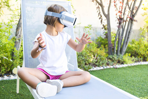 Little girl with virtual reality glasses, looking to the right, trying to touch something in a virtual way, sitting on a sun lounger in the garden of her house. Metaverse, VR, game, digital concept. - Photo, image