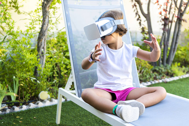 Little girl with virtual reality glasses, looking to the left, sitting on a sun lounger in the garden of her house. Metaverse, VR, game, digital and simulation concept. - Photo, Image