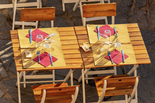 Close-up of an outdoor restaurant wooden table with paper tablecloth, cutlery, glasses and red paper napkins. View from above, Liguria, Italy, Europe. - Photo, image