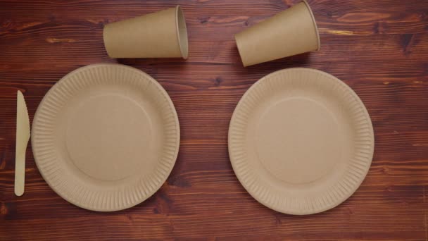 Paper cups, dishes, and wooden cutlery on wooden background. Eco craft paper tableware. Recycling or eco-friendly concept. Top view. - Filmati, video