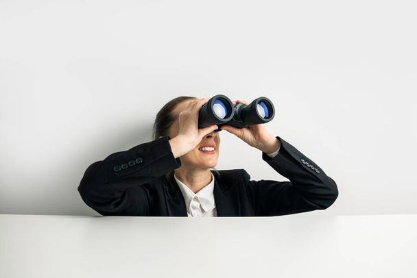 Business woman looking through binoculars peeking out from behind white table. - Valokuva, kuva