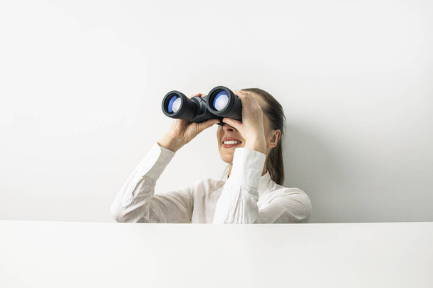 Smiling young woman in shirt looking through binoculars peeking out from behind cardboard. - Zdjęcie, obraz