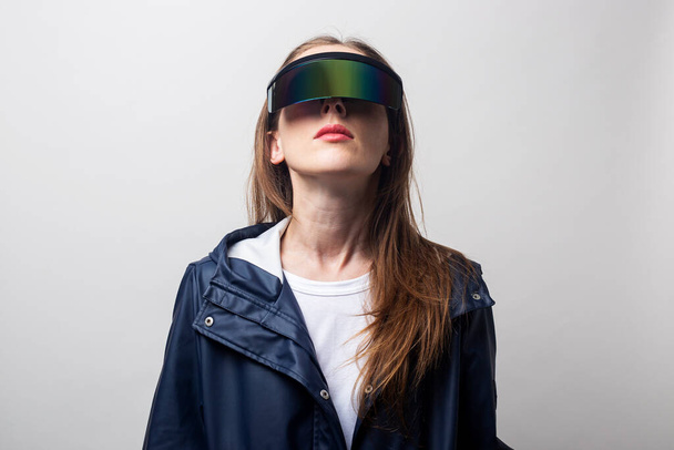 Young woman in virtual reality glasses in a blue jacket looks up on a light background.  - Photo, Image