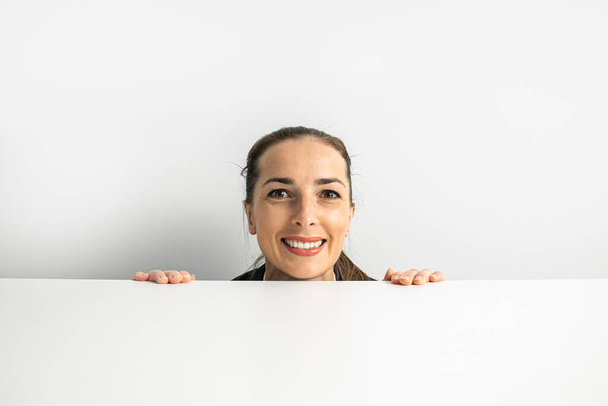 Smiling young woman peeking out from behind white table on white background. - 写真・画像