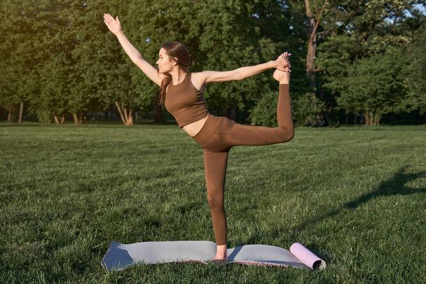 Una joven practica yoga en un parque de la ciudad mientras está de pie en una pose de guerrero. Deportes al aire libre. - Foto, imagen