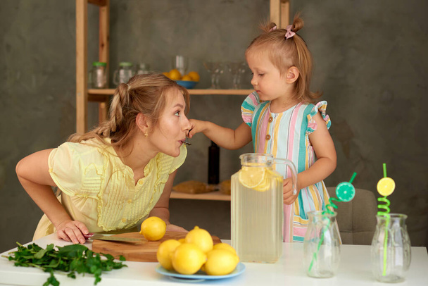 little daughter gives mom to taste homemade lemonade from spoon in kitchen - Foto, imagen