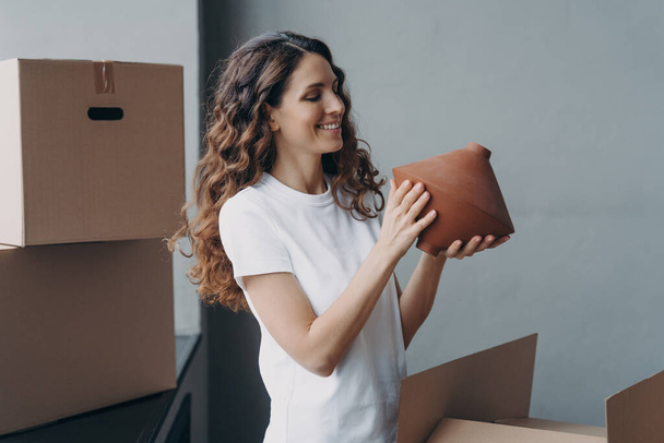 Relocation and delivery service concept. Happy girl unpacking cardboard boxes and holding the vase. Woman in casual outfit. Smiling attractive spanish woman packing boxes to move. - 写真・画像