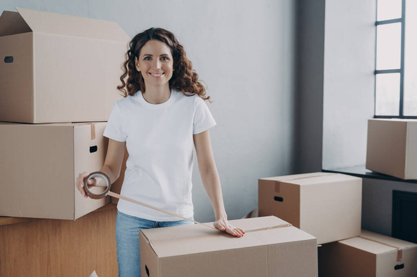 Young woman is loading and packing boxes for moving. European lady is wrapping cardboard boxes with packing tape. Moving service worker preparing boxes for shipping and storage. Delivery service. - Zdjęcie, obraz