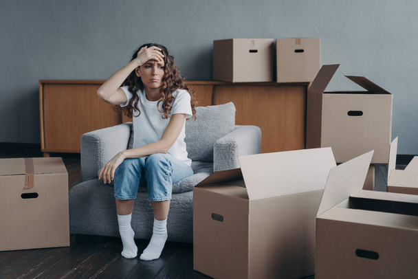 Headache from moving. Exhausted woman sitting among packed cardboard boxes. Spanish young lady in jeans and white t-shirt going to relocate. Depression and stress from moving concept. - Photo, image