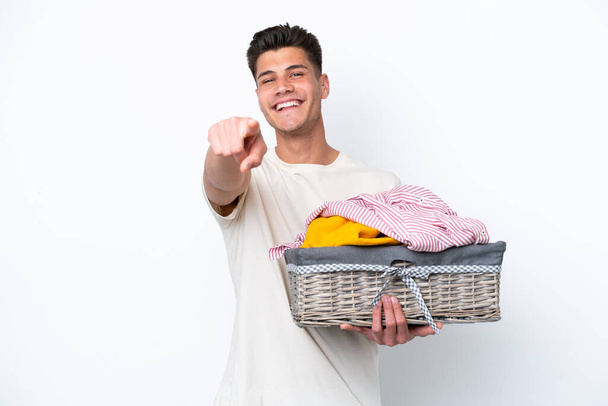 Joven hombre caucásico sosteniendo cesta de lavandería aislado sobre fondo blanco apuntando frente con expresión feliz - Foto, Imagen