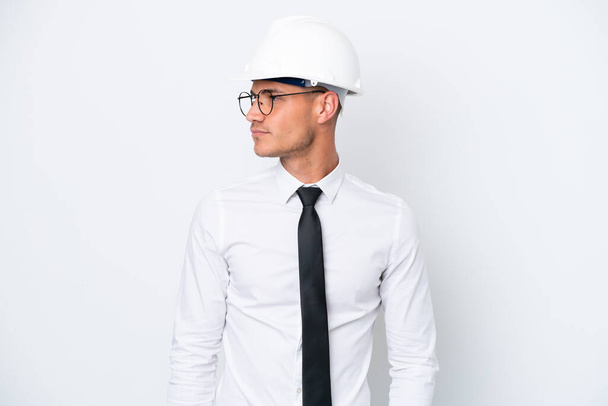 Giovane architetto uomo caucasico con casco e tenendo cianografie isolate su sfondo bianco guardando lateralmente - Foto, immagini