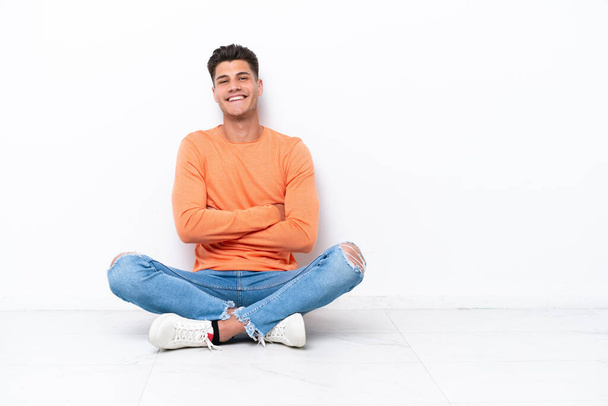 Young man sitting on the floor isolated on white background with arms crossed and looking forward - Fotografie, Obrázek
