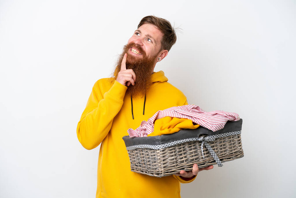 Rotschopf mit Bart hält einen Wäschekorb auf weißem Hintergrund und blickt lächelnd nach oben - Foto, Bild