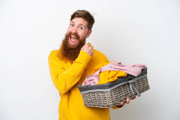 Redhead man with beard holding a clothes basket isolated on white background celebrating a victory - Photo, Image