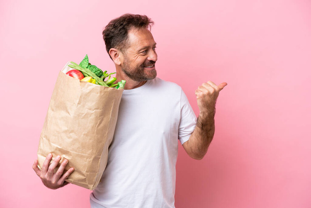 Middle age man holding a grocery shopping bag isolated on pink background pointing to the side to present a product - Foto, Bild