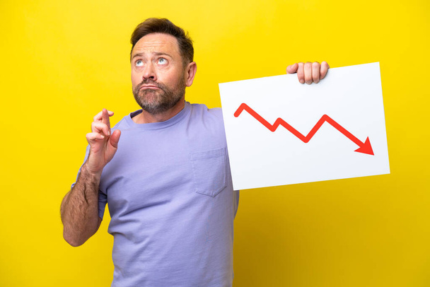 Middle age caucasian man isolated on yellow background holding a sign with a decreasing statistics arrow symbol with fingers crossing - Photo, Image