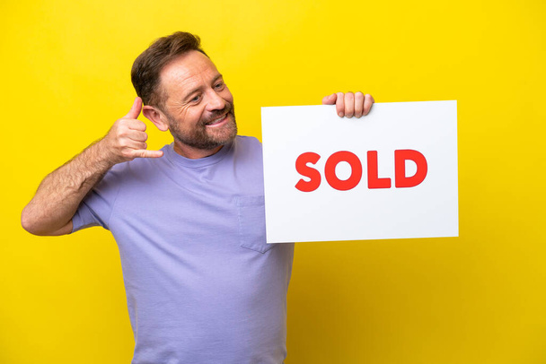 Middle age caucasian man isolated on yellow background holding a placard with text SOLD and doing coming gesture - Fotó, kép
