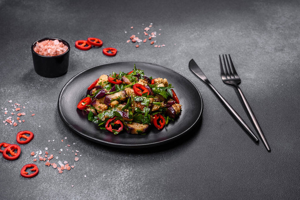 A salad of baked aubergine, sweet pepper, garlic, zucchini and parsley in a black plate against a dark concrete background. Vegetarian food - Photo, Image