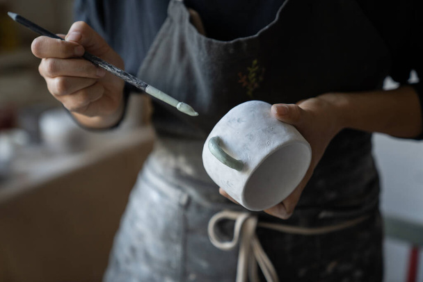 Experienced woman in black apron enjoys working with handmade craft in pottery studio. Female artisan uses brush to paint white ceramic cup with creative pattern standing on blurred background closeup - 写真・画像