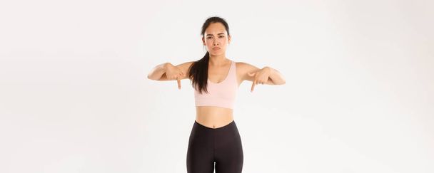 Sport, wellbeing and active lifestyle concept. Upset and disappointed asian sportswoman, woman in fitness clothing pointing fingers down with sulking face, complaining on banner, white background. - Photo, Image