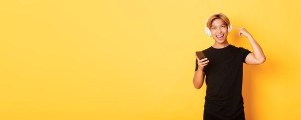 Šťastný spokojený asijský chlap má rád hudbu nebo podcast, ukazuje prstem na sluchátka, drží smartphone, stojící žluté pozadí. - Fotografie, Obrázek