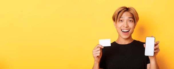 Erstaunlich glücklicher asiatischer Kerl zeigt Kreditkarte und Smartphone-Bildschirm, lächelt fasziniert, steht auf gelbem Hintergrund. - Foto, Bild