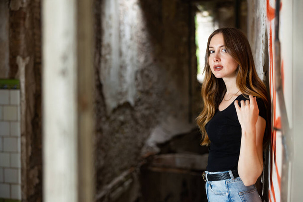 Portrait of attractive romantic young woman with flowing long hair in jeans and black tee shirt posing near brick wall in ruined building.. - Photo, Image