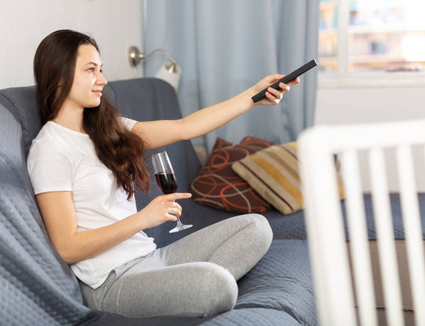 Positive young woman sitting on couch at home holding tv remote and glass of red wine - Foto, Imagem