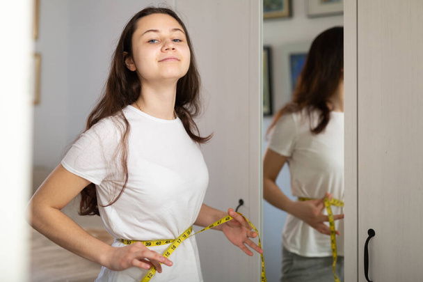 Retrato de una joven sonriente tomando medidas de su cintura usando cinta métrica de pie cerca del espejo en casa - Foto, Imagen