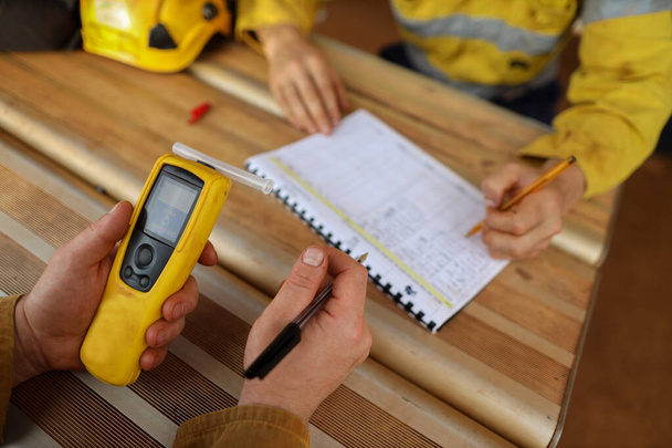 Safety workplace construction miner hand holding checking at breathalyzer breath alcohol testing equipment unit while his friend writing zero alcohol in the blood system on the daily sheet paper prior to work on construction mite site - Фото, изображение