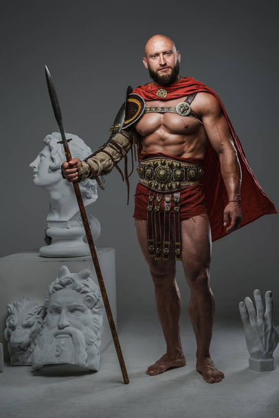 Portrait of greek gladiator from past holding spear posing around greek busts. - Photo, Image