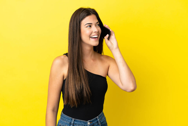 Young Brazilian woman isolated on yellow background keeping a conversation with the mobile phone - Photo, Image