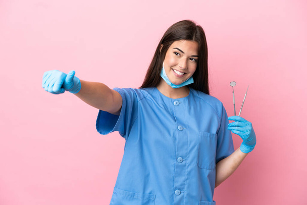 Mujer dentista sosteniendo herramientas aisladas sobre fondo rosa dando un gesto hacia arriba pulgares - Foto, Imagen